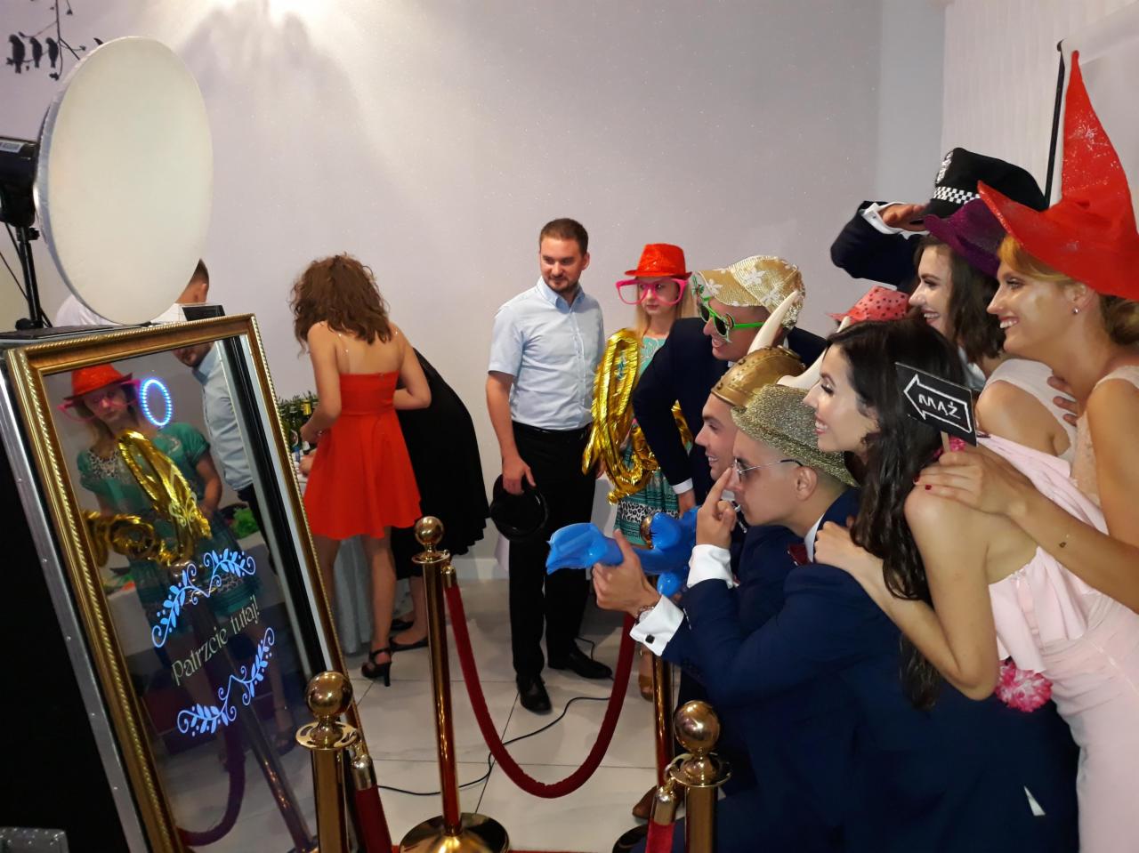 Grupa osób w przebraniach pozuje do zdjęcia w lustrzanej kabinie z napisem 'Patrzcie tutaj!', otoczonej złotymi słupkami oddzielającymi strefę fotografowania.