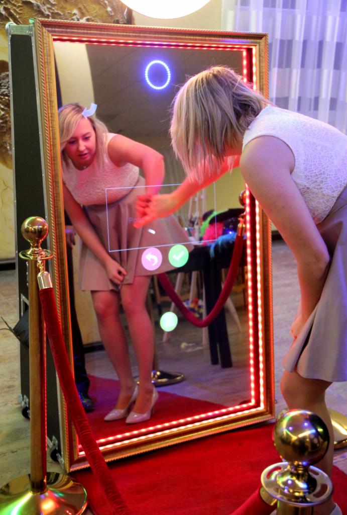 Kobieta w sukience używa fotobudki w kształcie lustra z interaktywnym ekranem dotykowym i czerwonym dywanem.