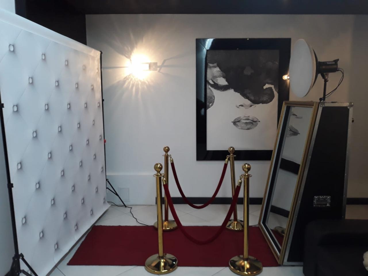 Elegancka aranżacja z białą pikowaną ścianką, złotymi słupkami z czerwonym sznurem, lustrem, lampą studyjną i czarno-białym portretem na ścianie, tworząca strefę zdjęć.