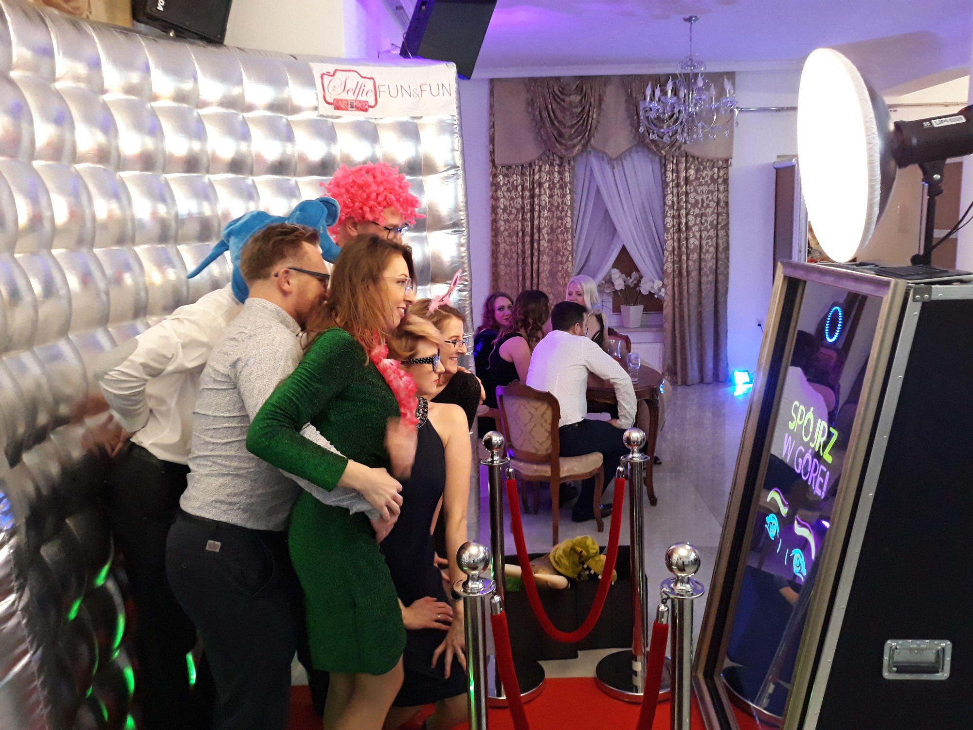 Grupa osób w przebraniach pozuje do zdjęcia w dmuchanej srebrnej kabinie z napisem 'Selfie FUN&FUN', w tle widoczny stolik z gośćmi i dekoracje sali.