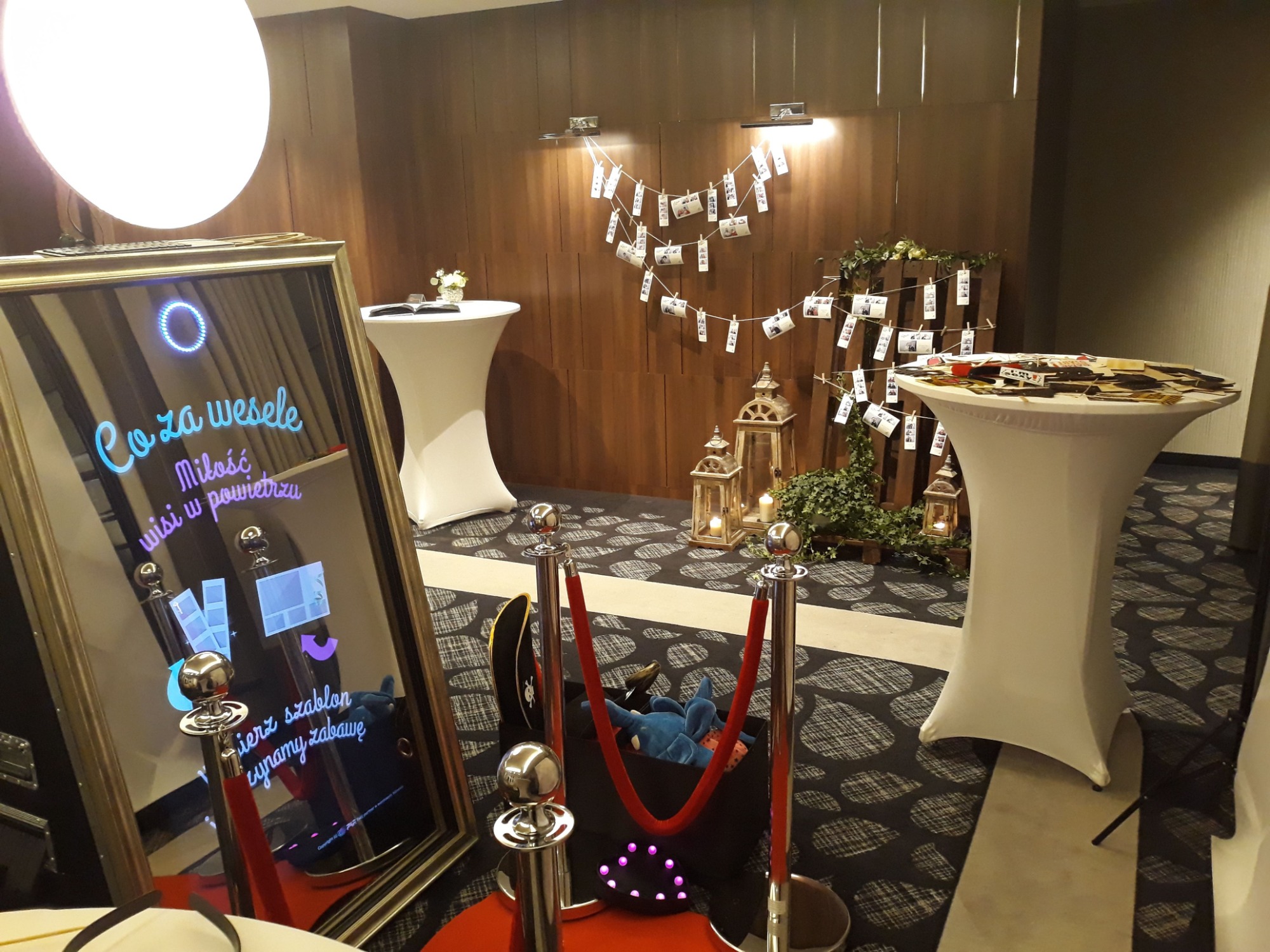 Fotobudka w lustrzanej ramie z wyświetlanym tekstem 'Co za wesele, miłość wisi w powietrzu' na tle dekoracji weselnej z lampionami, zdjęciami na sznurkach i rekwizytami na stoliku.
