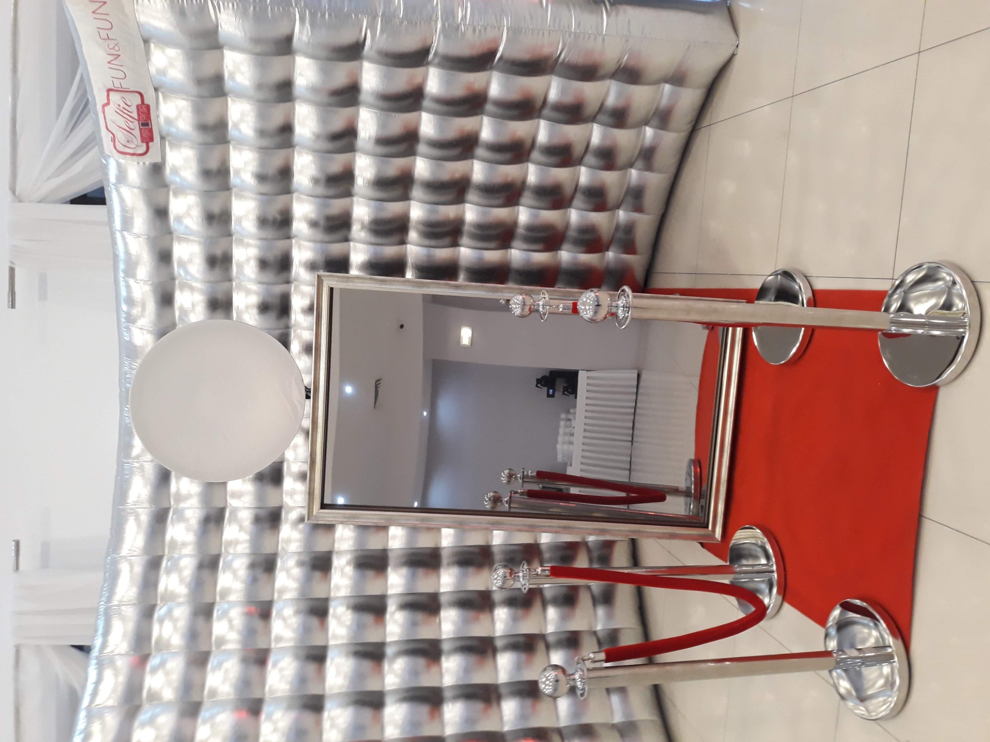 Srebrna, dmuchana ścianka z logo 'Selfie Fun&Fun', lustro w srebrnej ramie na czerwonym dywanie z chromowanymi słupkami i czerwonymi sznurami.