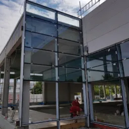 Montaż szklanej fasady budynku komercyjnego, widoczni pracownicy w czerwonych koszulkach, fragment elewacji z betonu architektonicznego, drabina oparta o budynek.