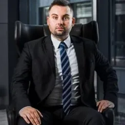 Portret prawnika siedzącego w skórzanym fotelu na tle okna, ubrany w czarny garnitur i niebiesko-biały krawat z klamrą.