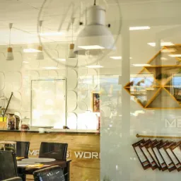 Wnętrze kawiarni z drewnianym blatem baru, białymi lampami wiszącymi i dekoracyjną ścianą 3D, widziane przez przeszkloną witrynę z menu i żółtymi półkami.