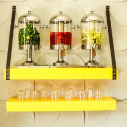 Trzy szklane dystrybutory napojów z kranikami, na żółtych półkach zawieszonych na skórzanych pasach, ustawione na tle ściany z dekoracyjnymi panelami.