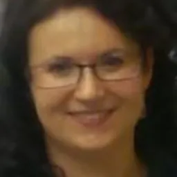 Portret uśmiechniętej kobiety w okularach, z ciemnymi kręconymi włosami, patrzącej wprost w obiektyw.