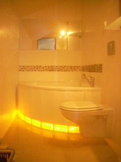 Oświetlona od dołu wanna narożna z hydromasażem i podwieszana toaleta w łazience wykończonej w jasnych odcieniach, z dekoracyjnym paskiem mozaiki.