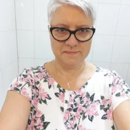 Portret kobiety w okularach i kwiecistej bluzce na tle białych płytek. Srebrne włosy, delikatny uśmiech, srebrny łańcuszek z wisiorkiem. Ujęcie z bliska, naturalne światło.