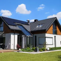 Nowoczesny dom jednorodzinny z panelami słonecznymi na dachu, podczas montażu przez pracownika w białej koszulce, błękitne niebo z chmurami w tle.