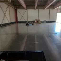 Wnętrze hali w trakcie budowy z widocznymi bloczkami, narzędziami i osobą leżącą na podłodze w oddali, metalowa konstrukcja dachu i białe ściany.