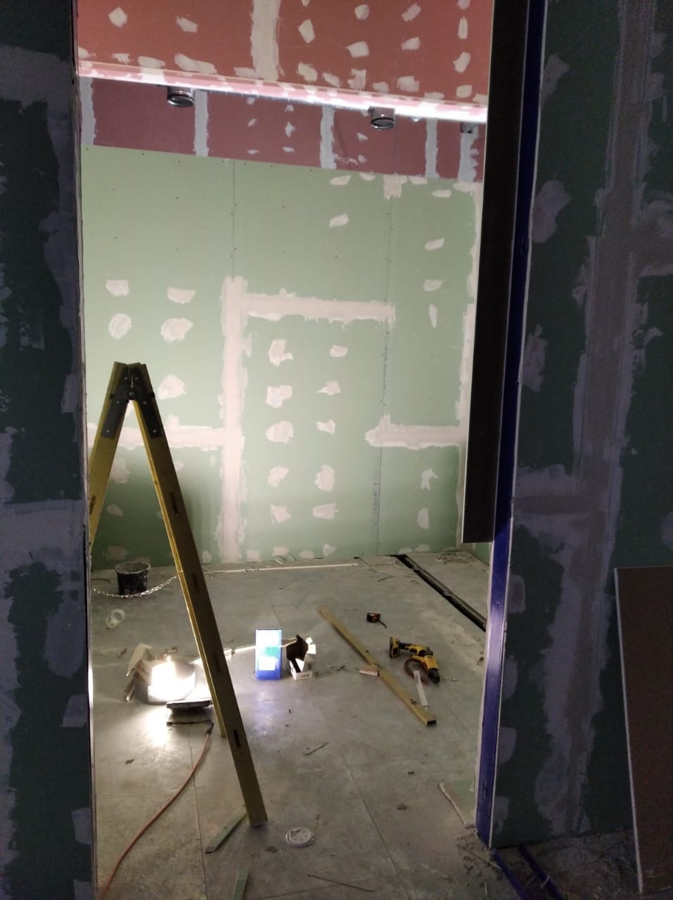 Wnętrze w trakcie prac wykończeniowych: ściany z płyt gipsowo-kartonowych z widocznymi spoinami, drabina, narzędzia i lampa oświetlająca podłogę.