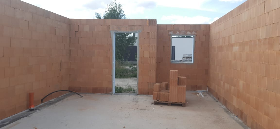 Wnętrze w stanie surowym otwartym z czerwonej cegły, widoczne otwory okienne i drzwiowe, paleta z cegłami w środku pomieszczenia, instalacja elektryczna w peszlu przy podłodze, widok na zieleń za...