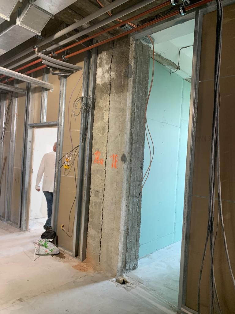 Wnętrze budynku w trakcie prac, widoczny surowy betonowy filar z pęknięciami, metalowa konstrukcja ścian działowych, instalacje pod sufitem i fragment ściany z płyt gipsowo-kartonowych. W tle...