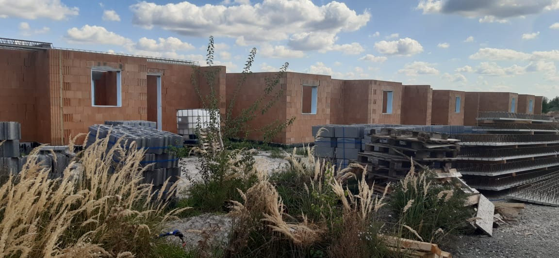 Wznoszone ściany parterowe z pustaków ceramicznych na budowie, widoczne otwory okienne i drzwiowe, palety z materiałami budowlanymi na pierwszym planie, w tle błękitne niebo z chmurami.