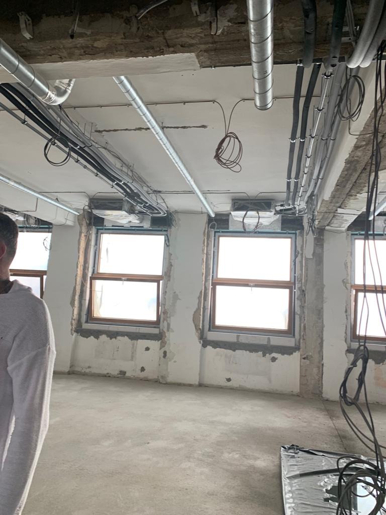 Wnętrze w stanie surowym: betonowa podłoga, okna z drewnianą ramą, ściany z resztkami tynku, widoczne instalacje pod sufitem, w tym rury wentylacyjne i kable.