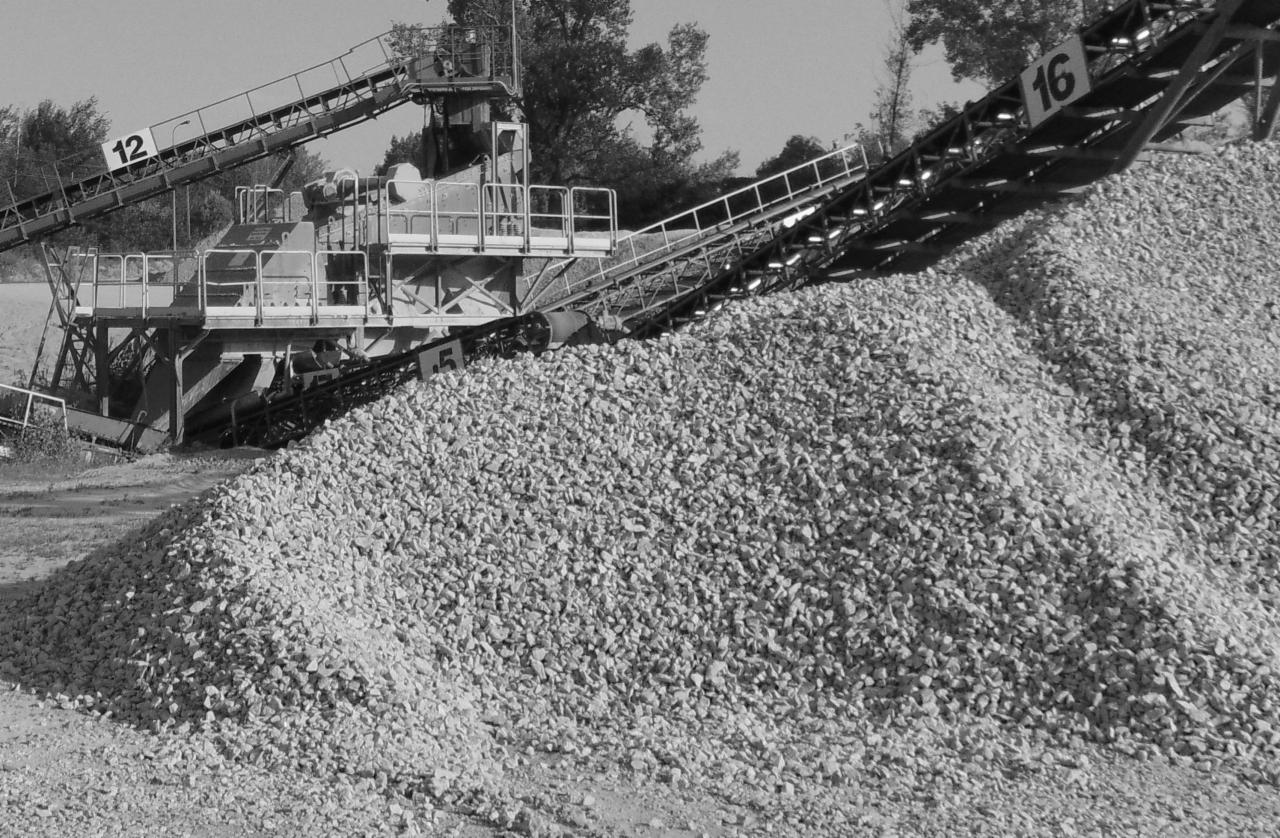 Czarno-białe zdjęcie hałdy drobnego, białego kruszywa wysypywanego z taśmociągu maszyny sortującej na tle drzew.