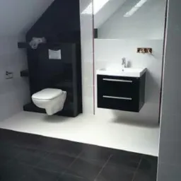 Nowoczesna łazienka na poddaszu z czarno-białymi płytkami, wiszącą toaletą i szafką pod umywalką w kolorze czarnym, oświetlona światłem dziennym z okna dachowego.