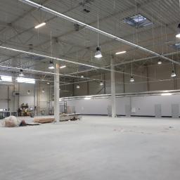 Voltek - Wnętrze hali przemysłowej z widoczną konstrukcją dachu, oświetleniem wiszącym i pracownikami na podnośniku koszowym przy ścianie, przygotowywanej do użytku.