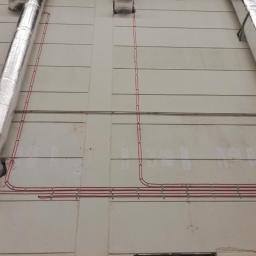 Voltek - Czerwone przewody elektryczne poprowadzone po zewnętrznej ścianie budynku, zabezpieczone metalowymi uchwytami, obok rur wentylacyjnych w srebrnej izolacji.