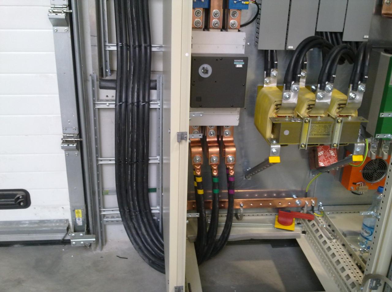 Rozdzielnica elektryczna z widocznymi kablami, transformatorami i elementami zabezpieczeń, zamontowana w pomieszczeniu technicznym.