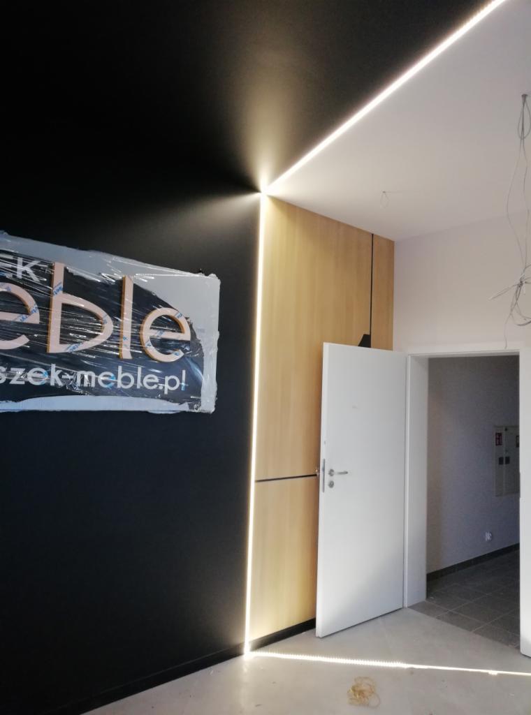 Nowoczesne oświetlenie LED wzdłuż ściany i sufitu w pomieszczeniu z czarną ścianą i drewnianą okładziną, widoczne logo firmy meblarskiej pod folią ochronną.