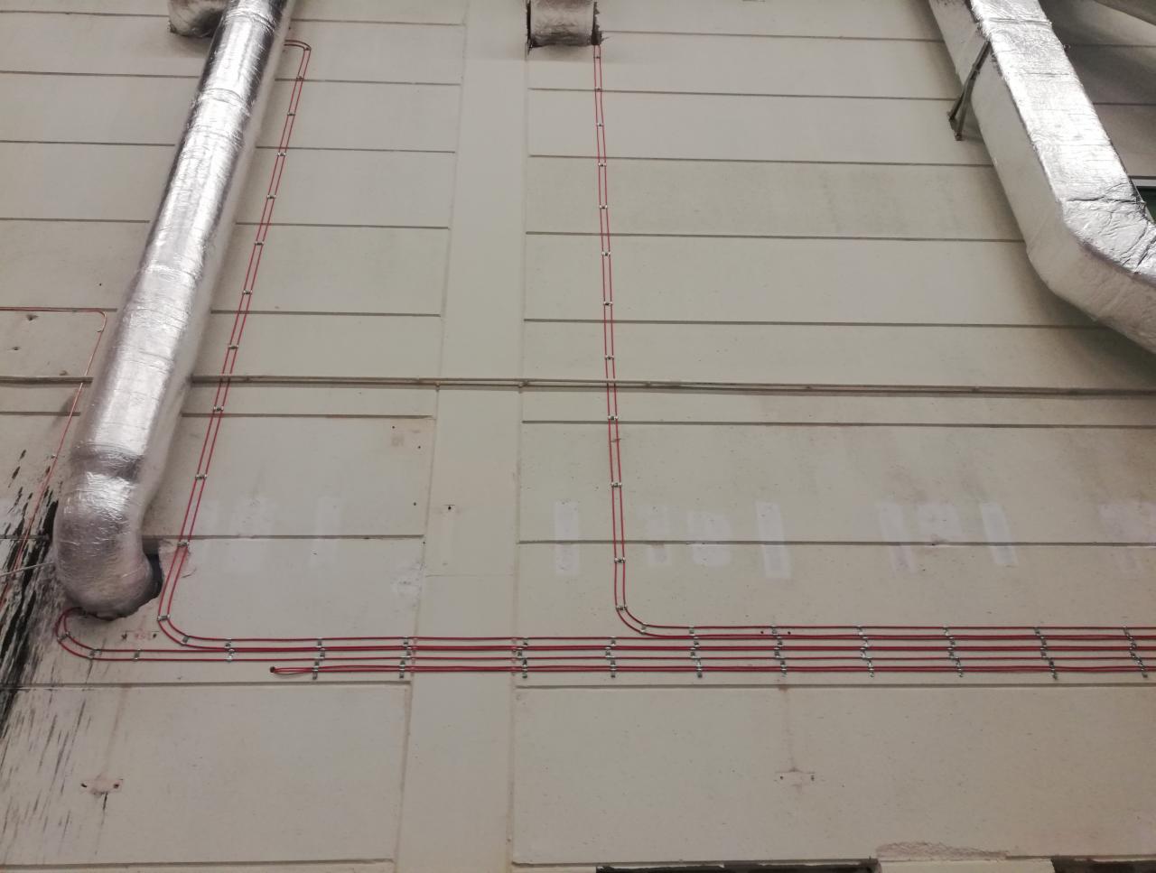 Czerwone przewody elektryczne poprowadzone po zewnętrznej ścianie budynku, zabezpieczone metalowymi uchwytami, obok rur wentylacyjnych w srebrnej izolacji.