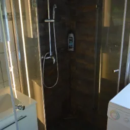 Nowoczesna kabina prysznicowa z deszczownicą i armaturą w kolorze chromu, brązowe płytki imitujące kamień, biała umywalka z szafką i pralka w tle.