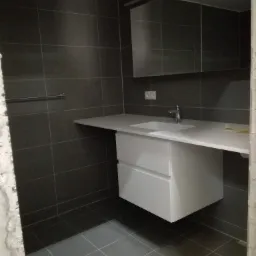 Nowoczesna łazienka w trakcie wykończenia: ciemnoszare płytki, biała szafka z umywalką, lustro szafkowe, widoczne ślady prac remontowych na ścianie.