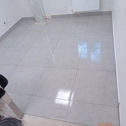 Caspol - Jasne, kwadratowe płytki podłogowe z szarą fugą, ułożone w nowym pomieszczeniu. Widoczne fragmenty instalacji i przygotowania do dalszych prac wykończeniowych.
