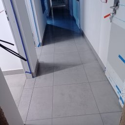 Caspol - Wyłożona szarymi płytkami podłoga w korytarzu, widok z perspektywy. Zabezpieczone folią i taśmą drzwi po obu stronach. Widoczny fragment progu.