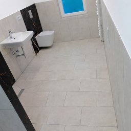 Caspol - Jasna łazienka po remoncie: beżowe płytki podłogowe i ścienne, wisząca toaleta, umywalka na stelażu, dekoracyjny panel z czarnego marmuru. Błękitne okno.