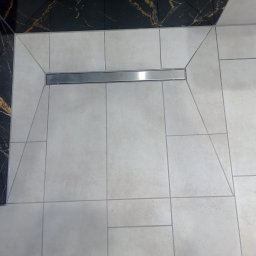 Caspol - Ułożone jasne płytki podłogowe z odpływem liniowym w kabinie prysznicowej. Fragment ściany wykończony czarnym marmurem. Geometryczny wzór płytek.