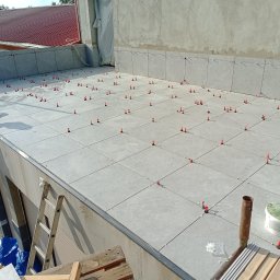 Caspol - Układanie szarych płytek na balkonie z użyciem systemu poziomowania. Widoczny glazurnik na drabinie i narzędzia pracy. Jasne światło słoneczne.