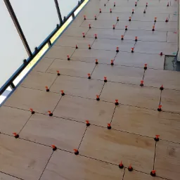 Układanie płytek na balkonie z użyciem pomarańczowych klinów poziomujących, widoczne fugi i fragment balustrady.