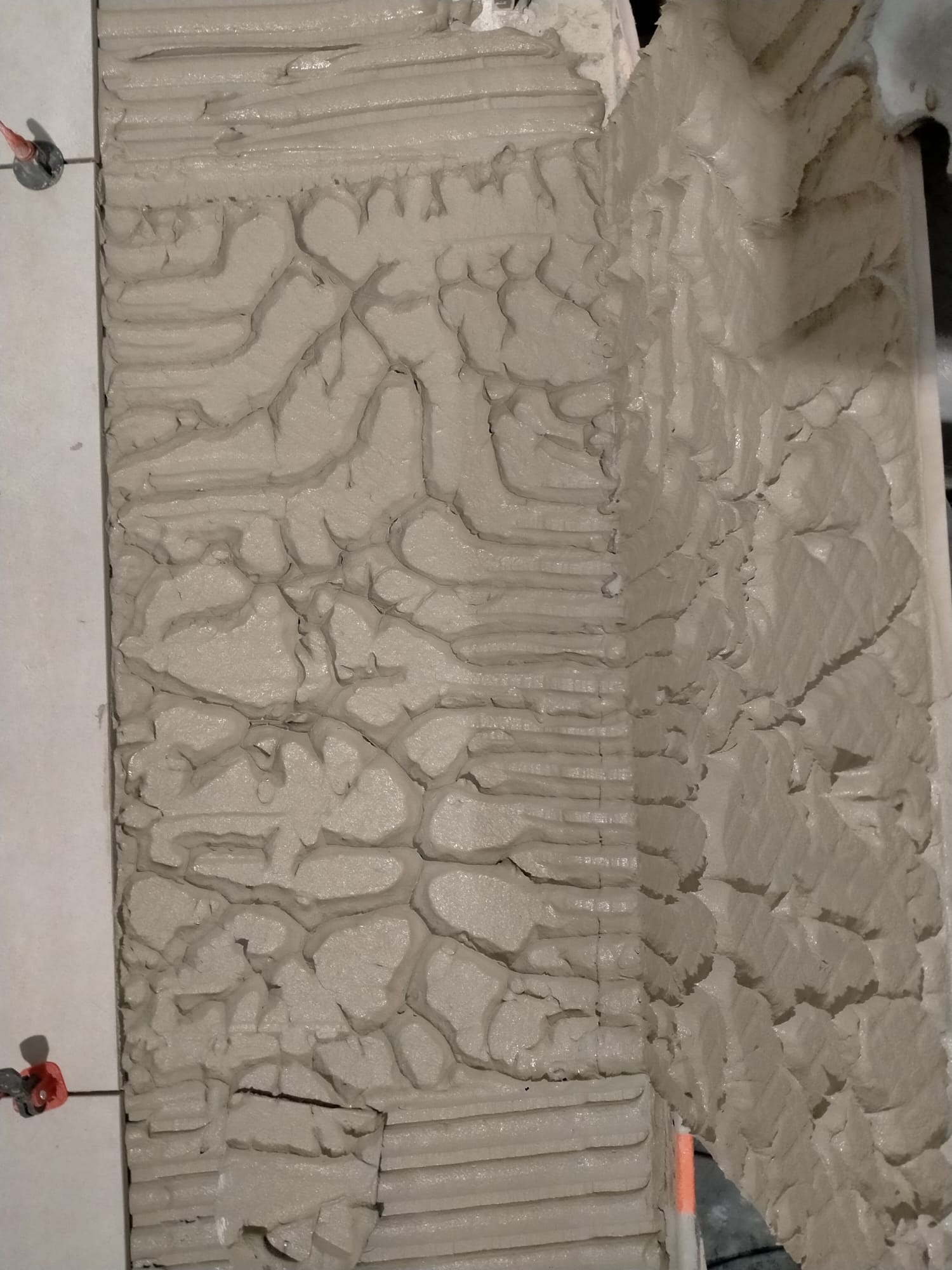 Świeżo nałożony klej na ścianie, przygotowany do układania płytek. Widoczne rowki i struktura kleju, a także fragment ułożonej płytki z krzyżykami dystansowymi.