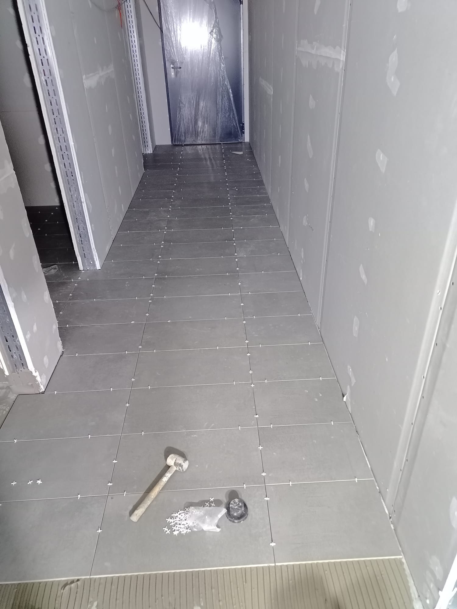 Układanie szarych płytek podłogowych w wąskim korytarzu. Widoczne krzyżyki dystansowe, młotek i odpływ podłogowy. Ściany wykończone płytami gipsowo-kartonowymi.
