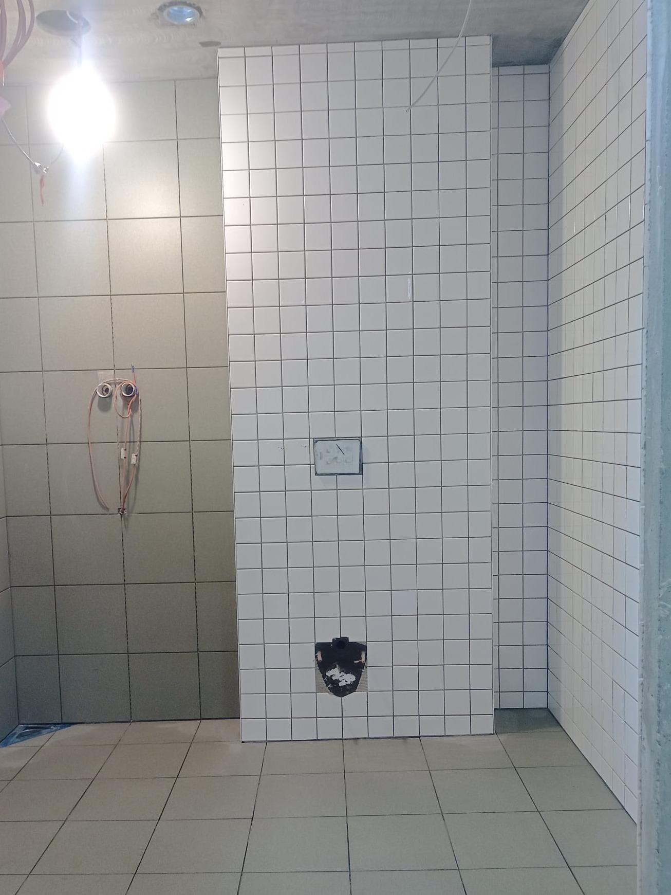 Wnętrze łazienki w trakcie prac wykończeniowych. Częściowo ułożone białe, kwadratowe płytki na ścianach i podłodze. Widoczne otwory na instalacje i wystające przewody.