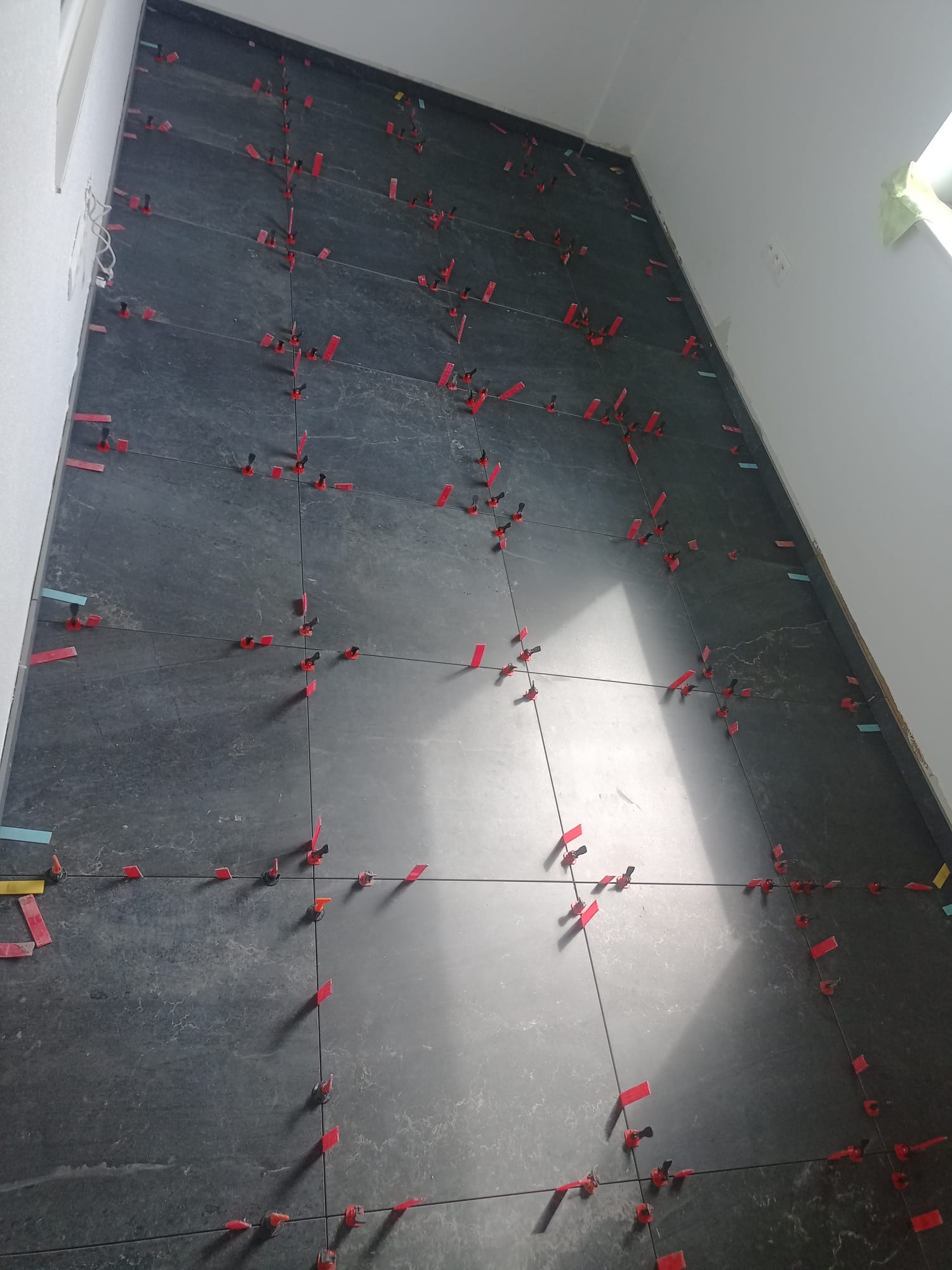 Układanie ciemnych, matowych płytek na ścianie z użyciem systemu poziomowania. Widoczne czerwone kliny i niebieskie elementy dystansowe. Świeżo położona glazura.