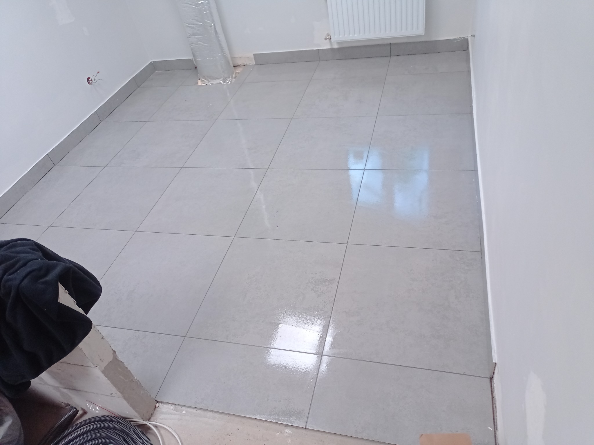 Jasne, kwadratowe płytki podłogowe z szarą fugą, ułożone w nowym pomieszczeniu. Widoczne fragmenty instalacji i przygotowania do dalszych prac wykończeniowych.