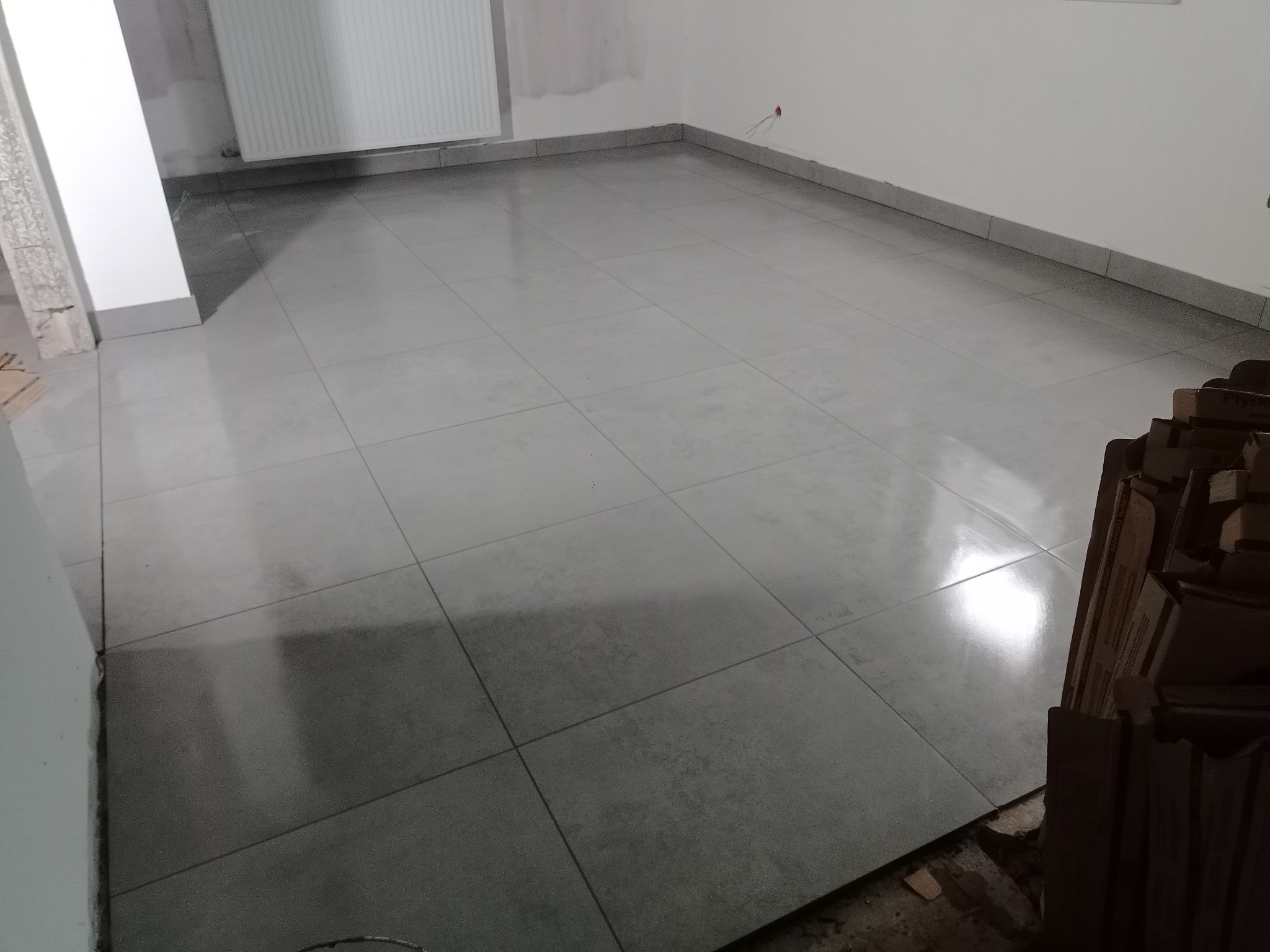 Wykończona podłoga w jasnoszarych, kwadratowych płytkach w trakcie remontu. Widoczne kartony z płytkami i fragment grzejnika. Minimalistyczny styl.