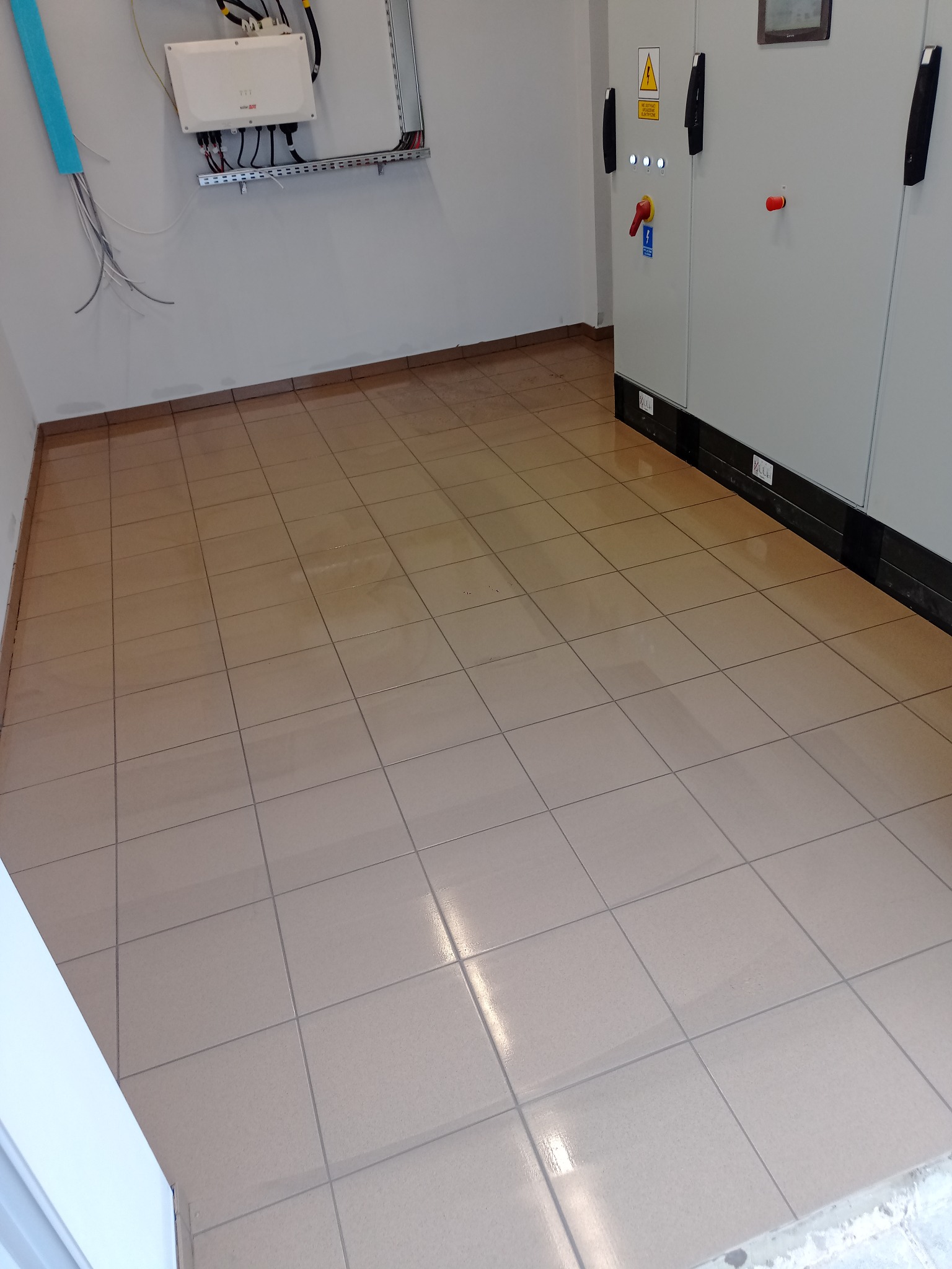 Beżowe płytki podłogowe w pomieszczeniu technicznym z szarą szafą sterowniczą i instalacją elektryczną na ścianie. Widok na świeżo wykafelkowaną podłogę.