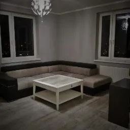 Szaro-beżowy narożnik z ciemnym obiciem i białym stolikiem kawowym z przeszklonym blatem na tle szarych ścian i nocnej panoramy miasta za oknami.