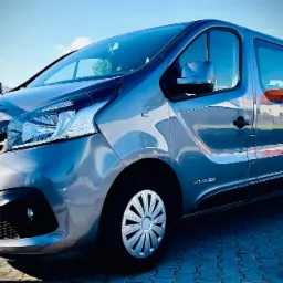 Szary bus Renault Trafic z aluminiowymi felgami, zaparkowany na kostce brukowej, widoczny fragment tablicy rejestracyjnej.