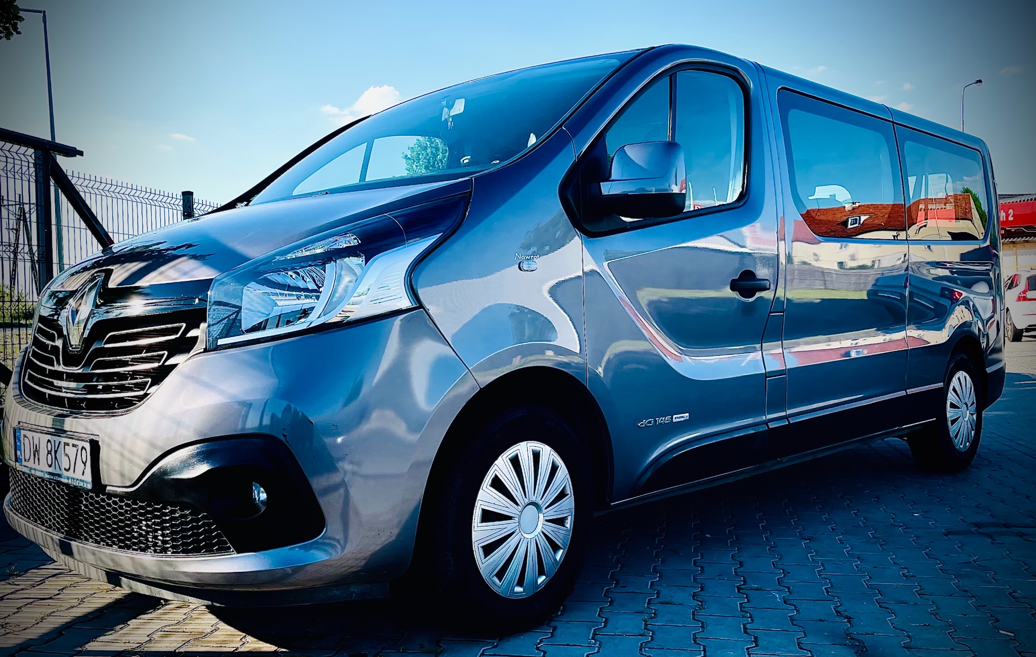 Szary bus Renault Trafic z aluminiowymi felgami, zaparkowany na kostce brukowej, widoczny fragment tablicy rejestracyjnej.