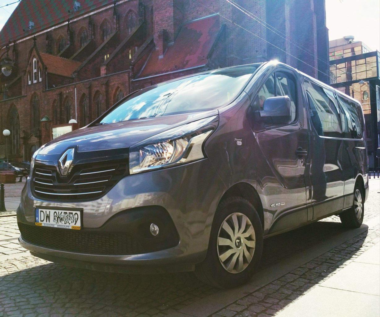 Szary van Renault Trafic zaparkowany na brukowanej ulicy z ceglanym budynkiem w tle, widoczna tablica rejestracyjna.