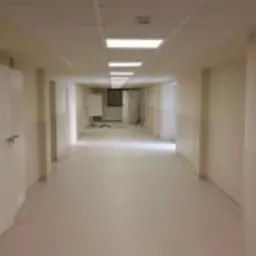 Długi korytarz w budynku biurowym z jasnymi ścianami, białymi drzwiami i podwieszanym sufitem z wbudowanym oświetleniem. Widoczne prace wykończeniowe w oddali, w tym niezabezpieczona ściana...