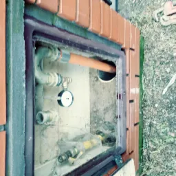 Studzienka rewizyjna z rurami, manometrem i zaworami, otoczona ceglaną obudową, widoczne narzędzia hydrauliczne na ziemi.