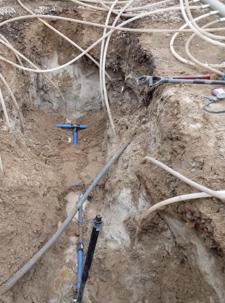 Wykop w ziemi z widocznymi rurami wodociągowymi i złączkami, narzędzia budowlane leżą na krawędzi wykopu.