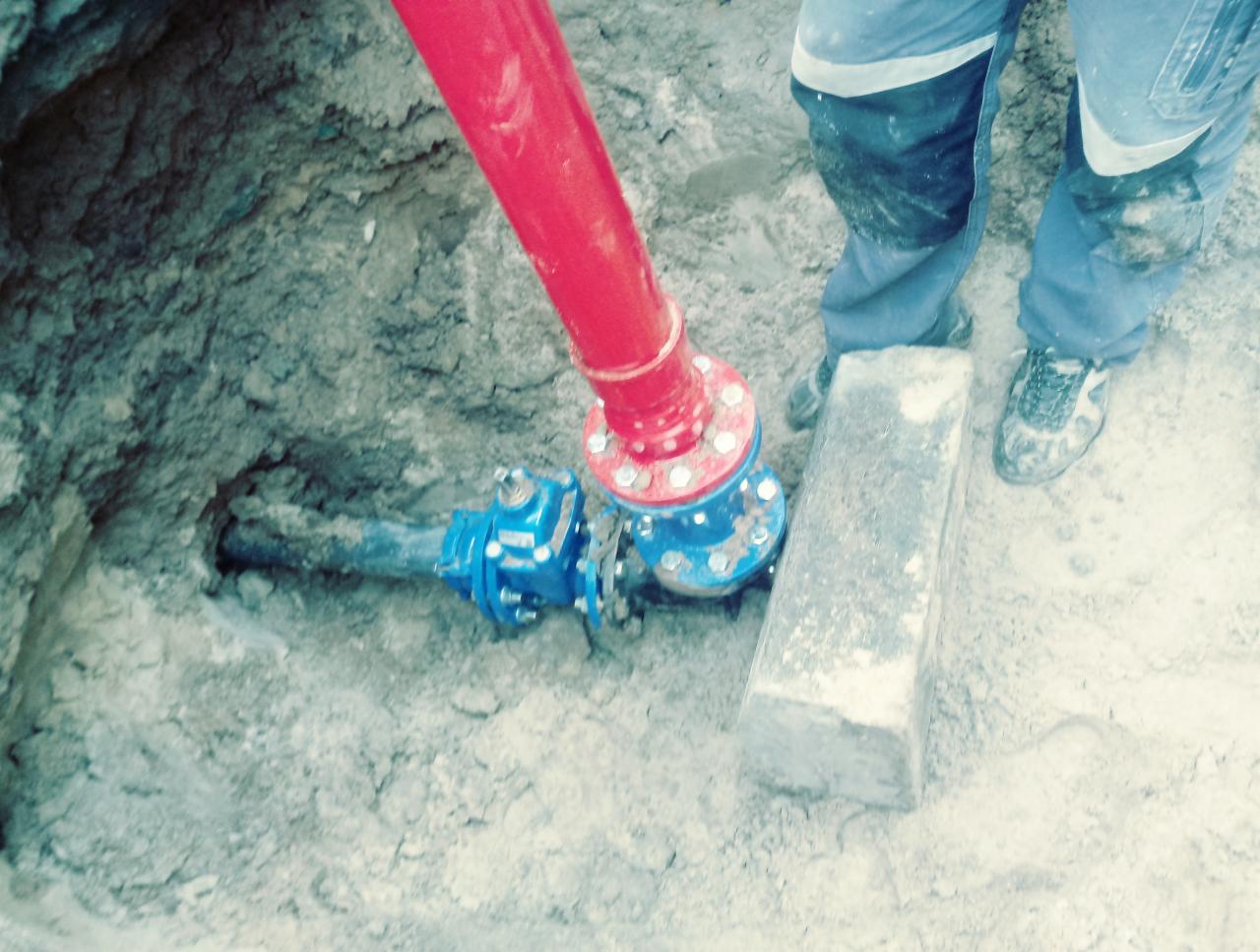 Wykonanie przyłącza wodociągowego: czerwona rura połączona z niebieską armaturą w wykopie ziemnym, widoczne buty pracownika i betonowa podpora.